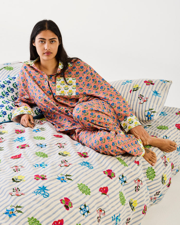 A woman wearing a pink floral pajama set with striped cuffs and a matching patterned duvet cover sits on a bed against a plain white wall.