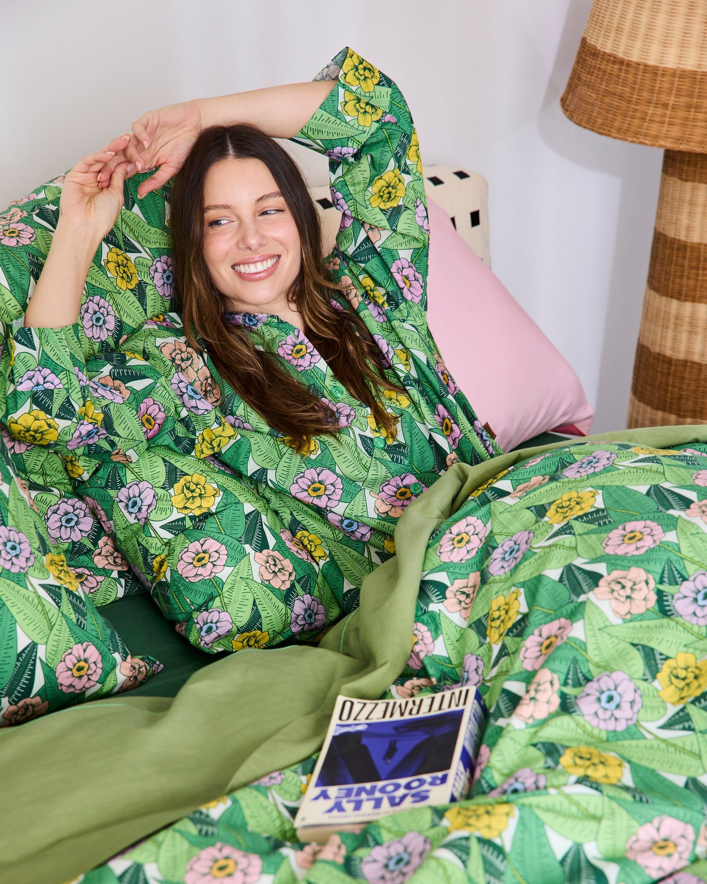 A woman reclining in bed wearing a green tropical floral robe and matching patterned bedding, with a pink pillow and a lamp visible in a bedroom setting.