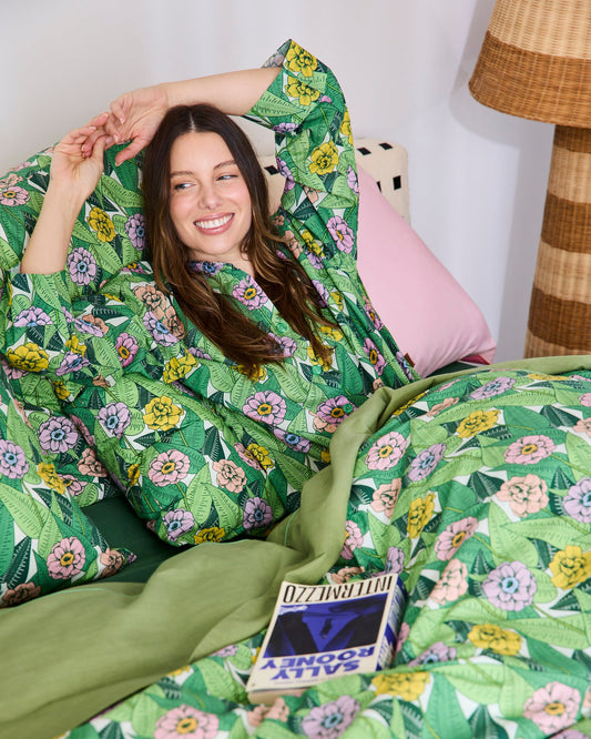 A woman reclining in bed wearing a green tropical floral robe and matching patterned bedding, with a pink pillow and a lamp visible in a bedroom setting.