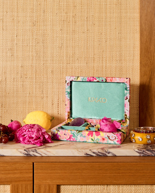 An open floral jewelry box with a mint green interior and gold 'RICO' lettering on the lid, styled on a marble-topped surface with fruit and a peony against a woven beige wall.