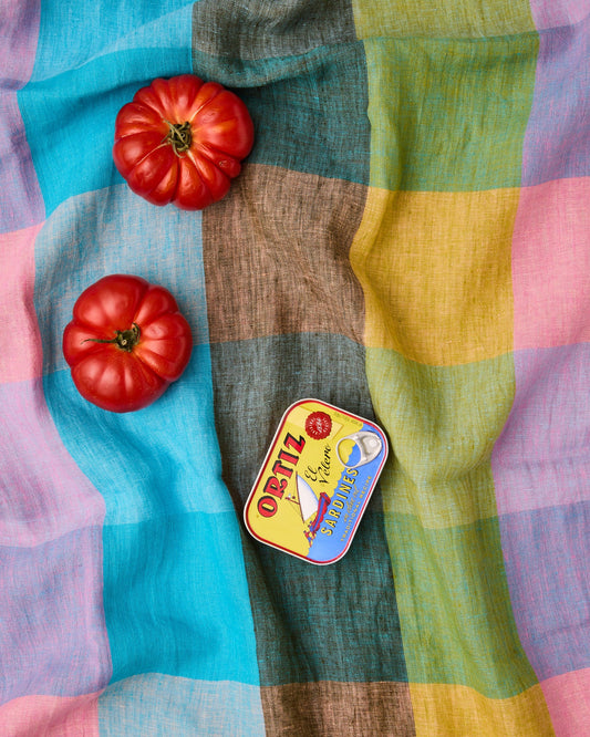 A colorful striped fabric with blocks of turquoise, brown, yellow, green, and pink, styled with two red tomatoes and a vintage Ostiz Serrameta tin on top.