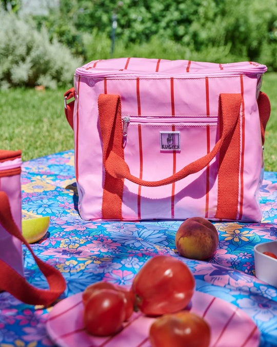 A pink cooler bag with red stripes and orange handles sits on a colorful floral picnic blanket outdoors on grass, with peaches and a small matching container nearby.