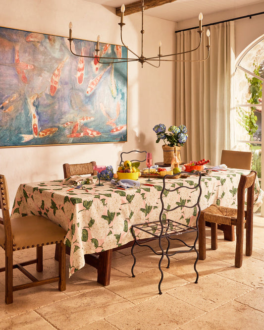 A dining room scene with a rectangular table covered in a cream tablecloth printed with green leaves and pink flowers. The table is set with fruit, flowers, and glassware, surrounded by wooden dining chairs.