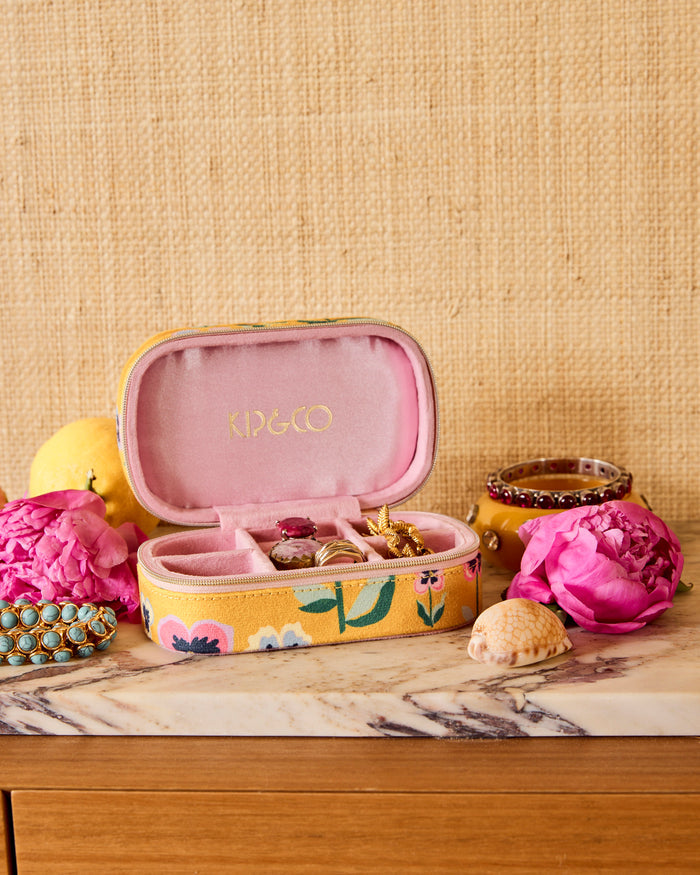A yellow floral jewelry box with a pink interior, open to display rings and earrings, styled on a marble surface with pink flowers and a woven beige backdrop.