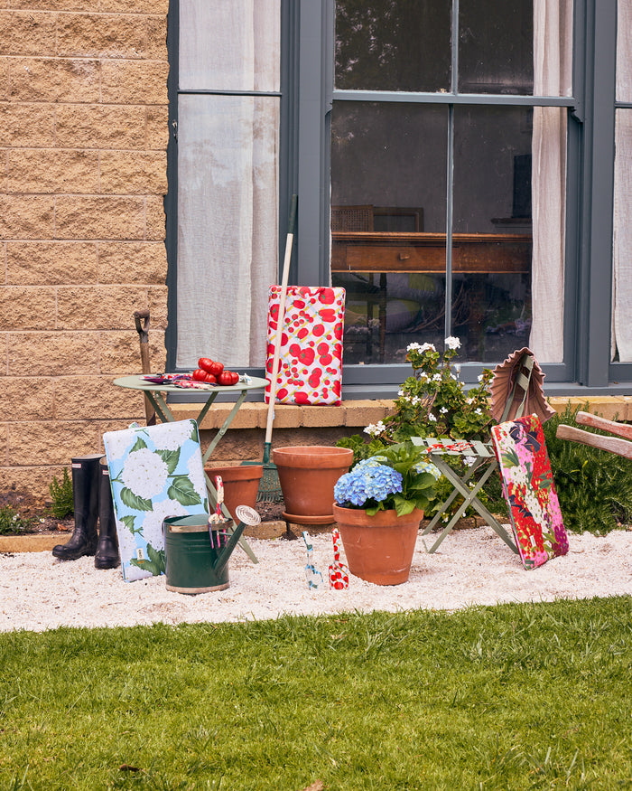 A styled outdoor garden scene in front of a house window, featuring terracotta pots, a small table, gardening tools, rain boots, and several fabric garden bags and cloths in colorful floral and leaf patterns.