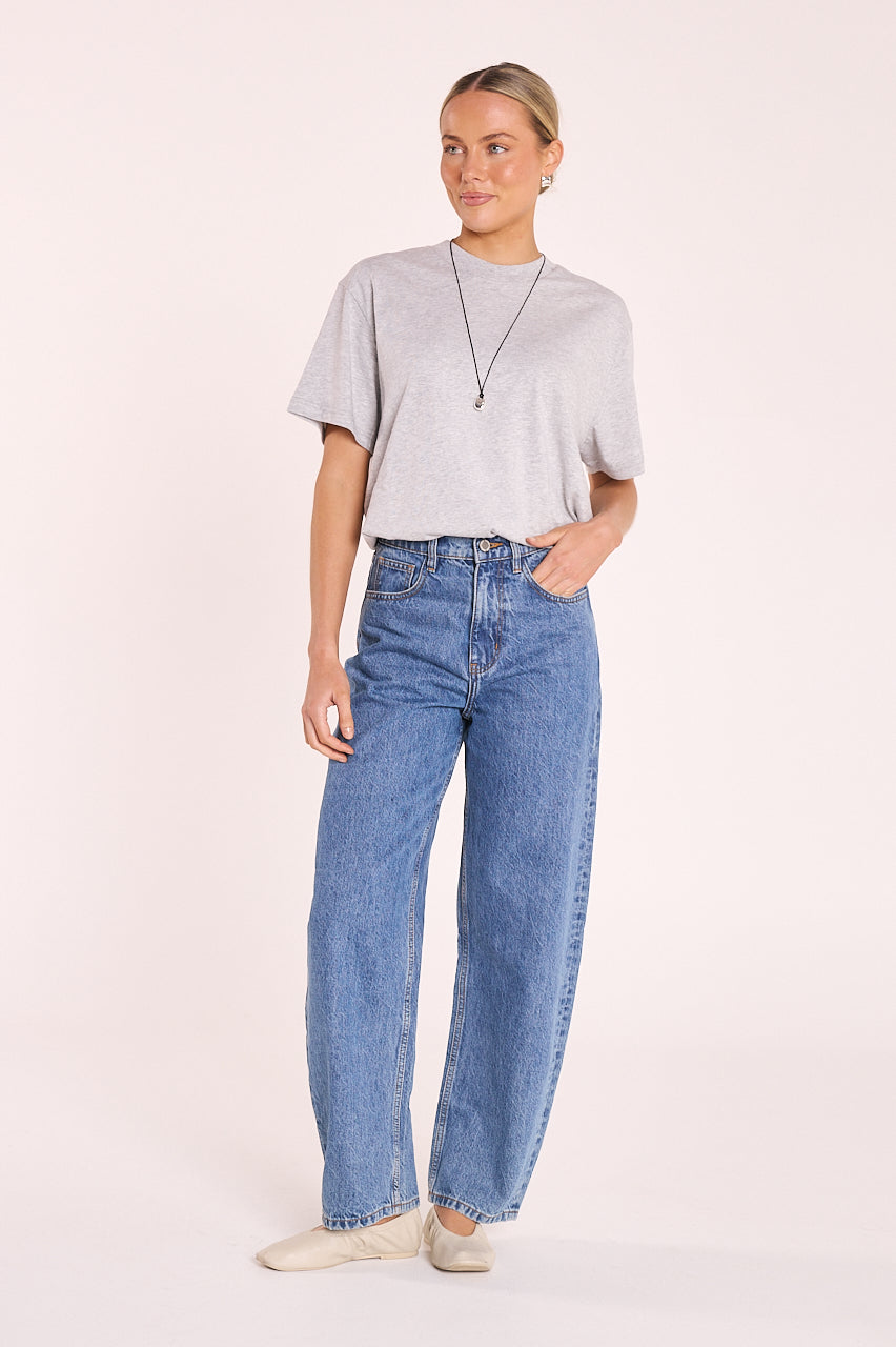A woman modeling a light gray short-sleeve t-shirt and high-waisted, wide-leg blue jeans against a solid pale pink studio background.