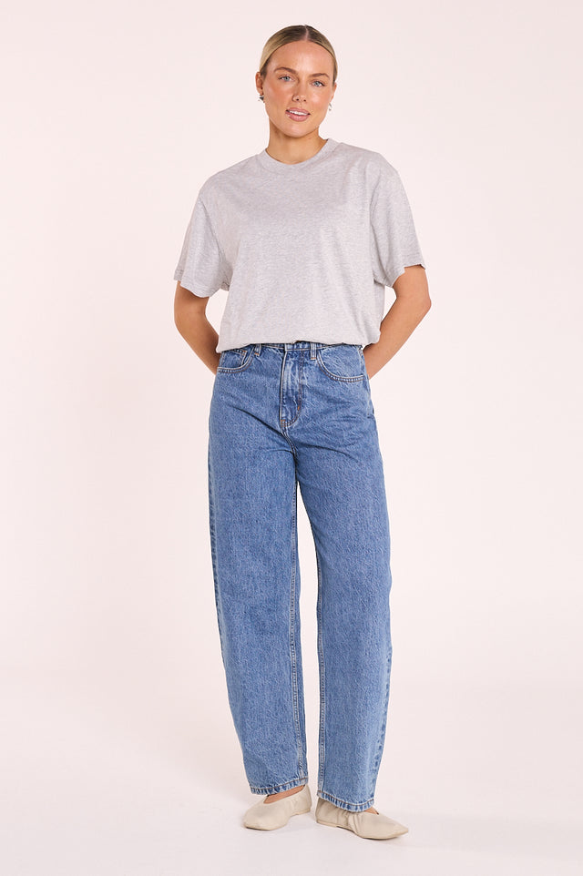 A woman modeling a light gray short-sleeve t-shirt and high-waisted, wide-leg blue jeans against a solid pale pink studio background.