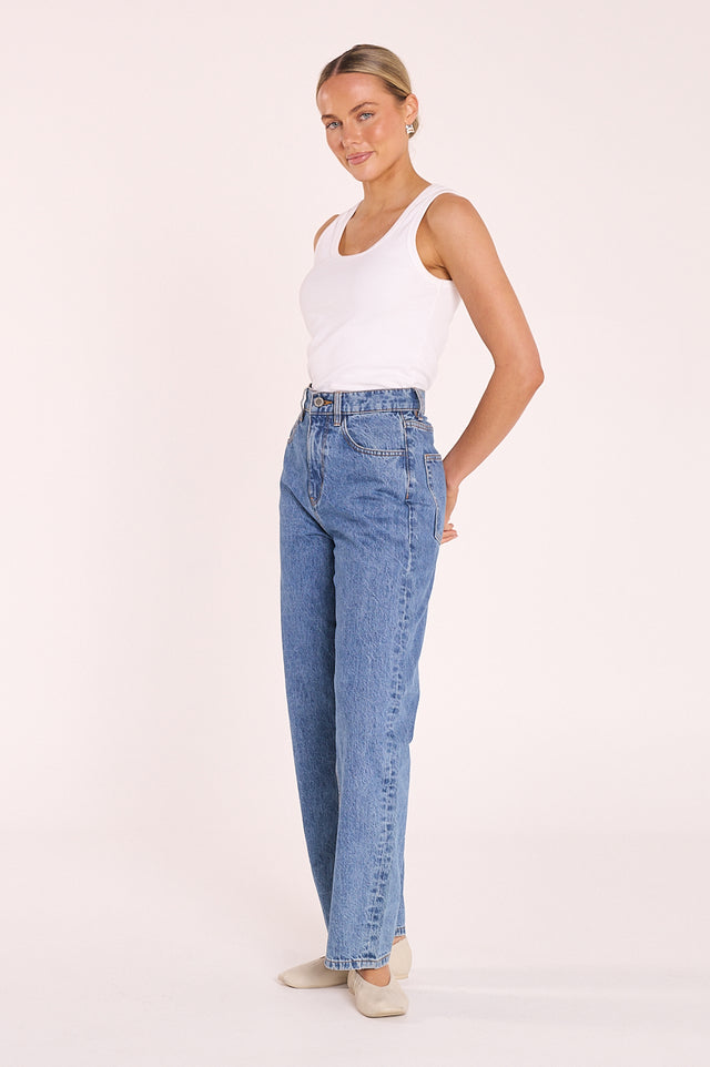 A woman modeling a pair of high-waisted, wide-leg blue jeans with a white tank top and beige shoes, standing against a solid light pink studio background.