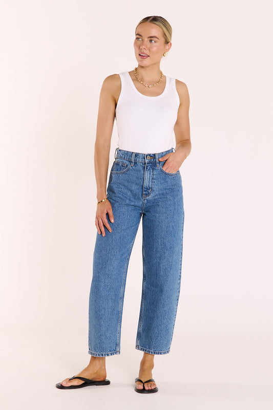 Woman wearing a white tank top and blue jeans on a light pink background
