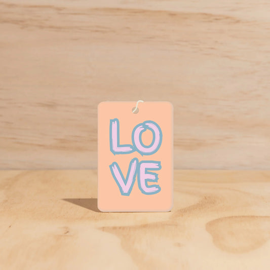 Rectangle car freshener with 'LOVE' text written on front in bold letters. Peach background with pink and blue lettering. 