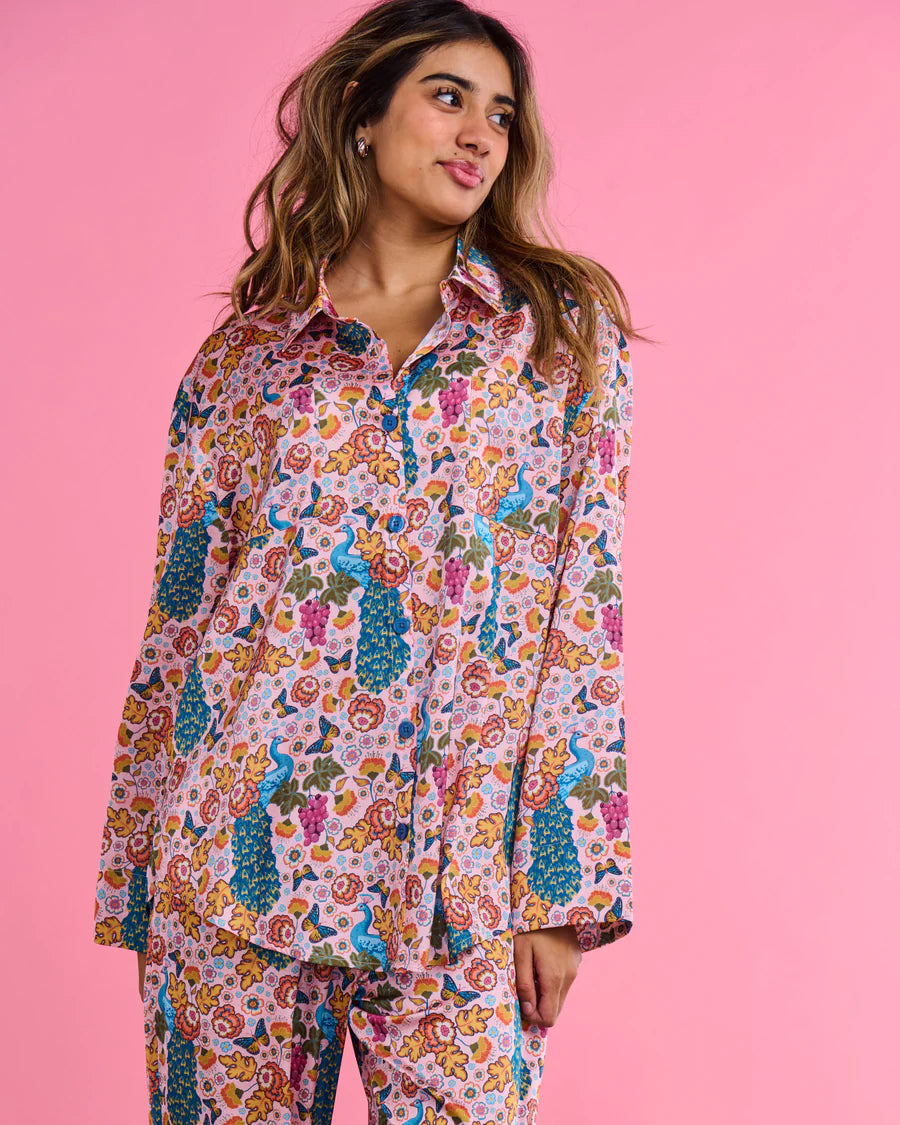 A woman modeling a matching pajama set with a colorful floral and peacock print, set against a solid pink background.