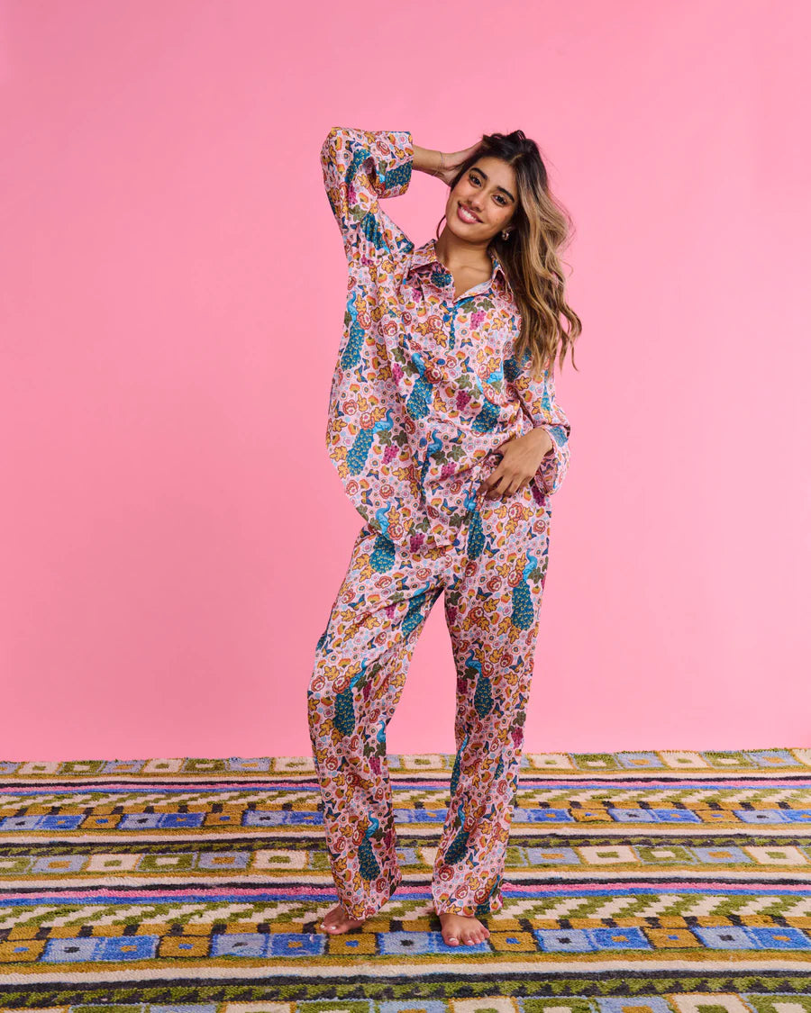 A woman modeling a matching two-piece pajama set with a colorful floral and paisley print, standing on a patterned rug against a solid pink background.