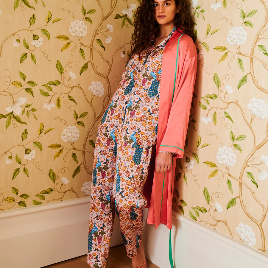 A woman stands barefoot in a room with floral wallpaper, wearing a matching pajama set with a multicolor floral print and a coral pink satin robe draped over one arm.