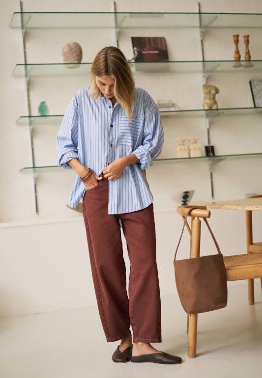 Woman wearing a striped shirt and brown pants in a room with shelves and a chair.