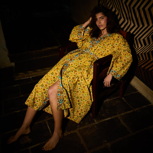 A woman reclining on a dark wooden chair in a dimly lit setting, wearing a yellow floral midi dress with blue and green accents.