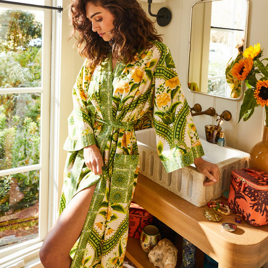 A woman wearing a green and yellow floral print kimono robe stands in a sunlit bathroom next to a sink, with a mirror, flowers, and toiletries visible in the background.
