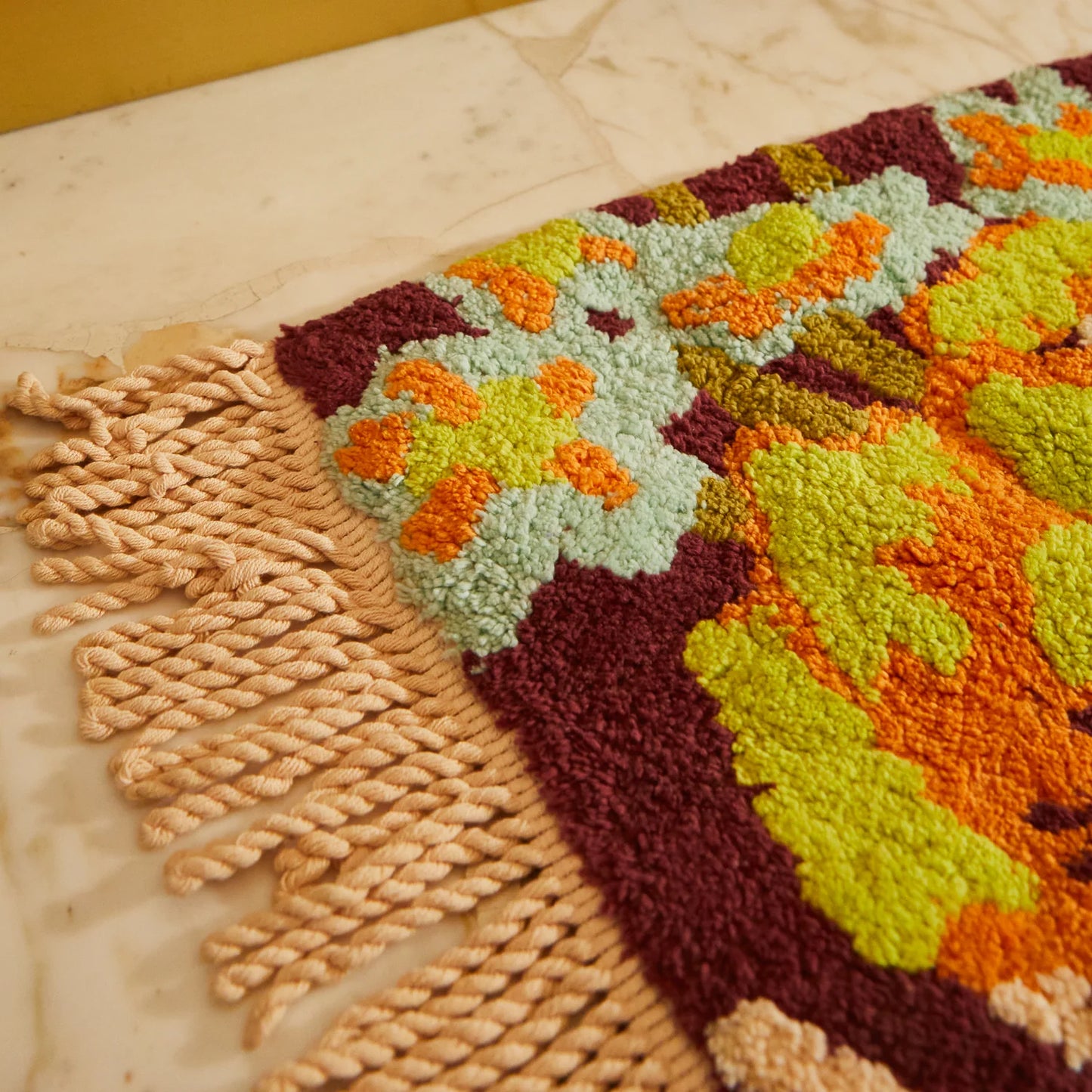 Close-up of a colorful woven bath mat with a cream tassel on a marble surface.