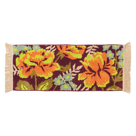 Floral patterned rug with orange and green flowers on a purple background, cream fringing along ends of bath runner. Photo has white background. 