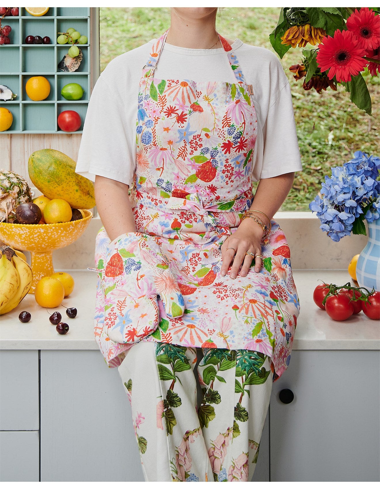 Person wearing a colorful apron and matching oven mitt in a kitchen setting with fruits and flowers. Photo showcases floral, colourful Meandering Meadow print. 