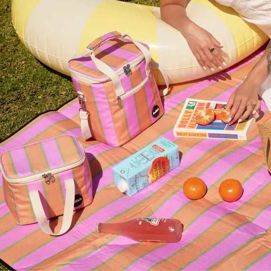 Colorful striped picnic blanket with bags, food items, and a magazine on a grassy area.