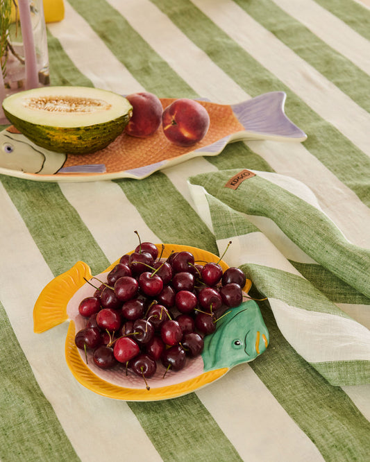 A styled tabletop scene with fish-shaped serving plates holding fruit, placed on a green-and-white striped cloth. The foreground plate holds dark red cherries, and the background plate holds a cut melon and two red fruits.