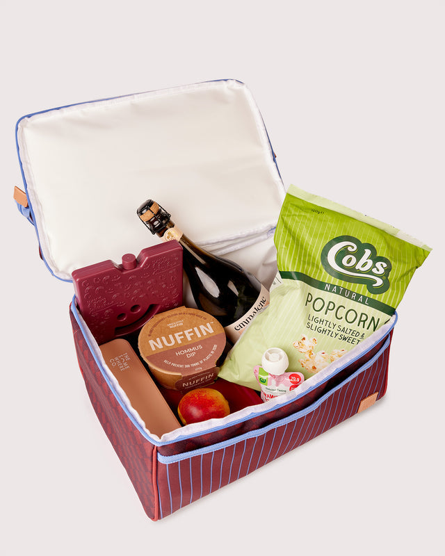 An open red picnic cooler with blue piping, filled with snacks and a bottle of champagne, photographed on a light gray background. Visible items include a bag of Cobs Natural Popcorn, a round Nuffin cheese container, a bottle, and a red wooden picnic plate.