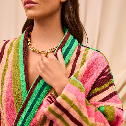 Woman stands wearing multicolour stripe robe and big gold accessories. Colours within robe include pink, green, burgundy and peach. 