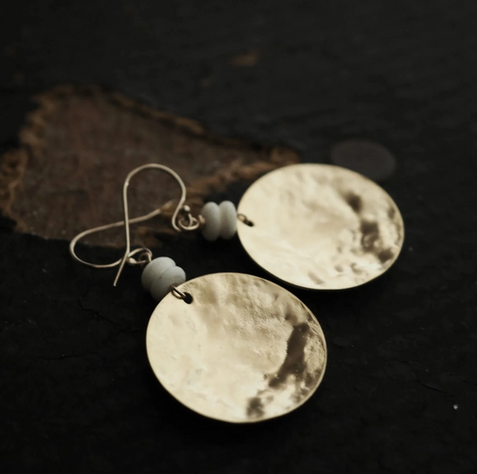 Pair of gold disc earrings, gold earwires and two white beads above circle disc pendant, with a dark background.