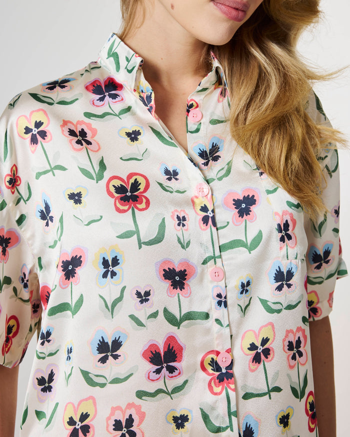 A close-up of a woman wearing a short-sleeve button-up shirt with a colorful floral print on a cream background. The print features pansy-like flowers in pink, red, blue, yellow, and black with green stems and leaves.