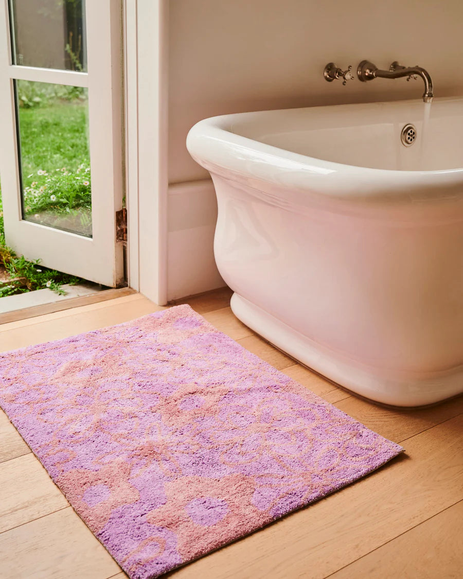 Purple bath mat in front of a white freestanding bathtub with a glass door leading to a garden.