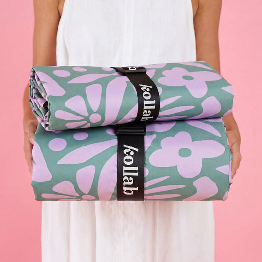 A person in a white sleeveless dress holds two rolled towels with a sage green and lavender floral pattern, secured by black straps with the brand name 'kollab' visible.