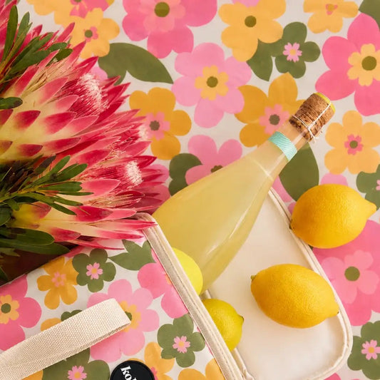 A styled flat-lay featuring a frosted glass bottle filled with pale yellow liquid, set on a white tray with lemons, against a vibrant pink, yellow, and green floral patterned backdrop with a large pink protea flower.