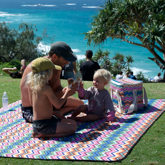 A family picnic scene on a colorful chevron-patterned blanket outdoors by the ocean, with a man, two children, a cooler, and a beach background.