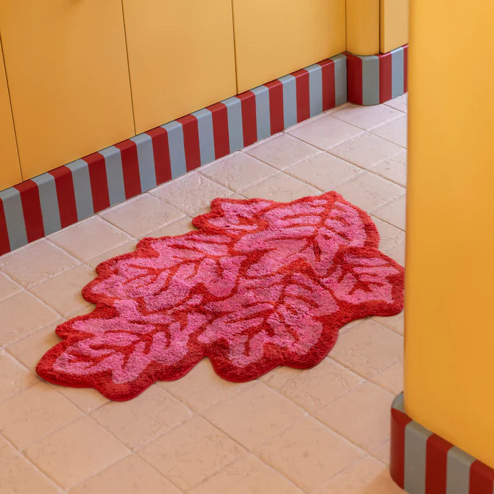 Pink and red doormat shaped like a multi leaf pattern on a tiled floor with yellow walls and red and gray striped baseboard.