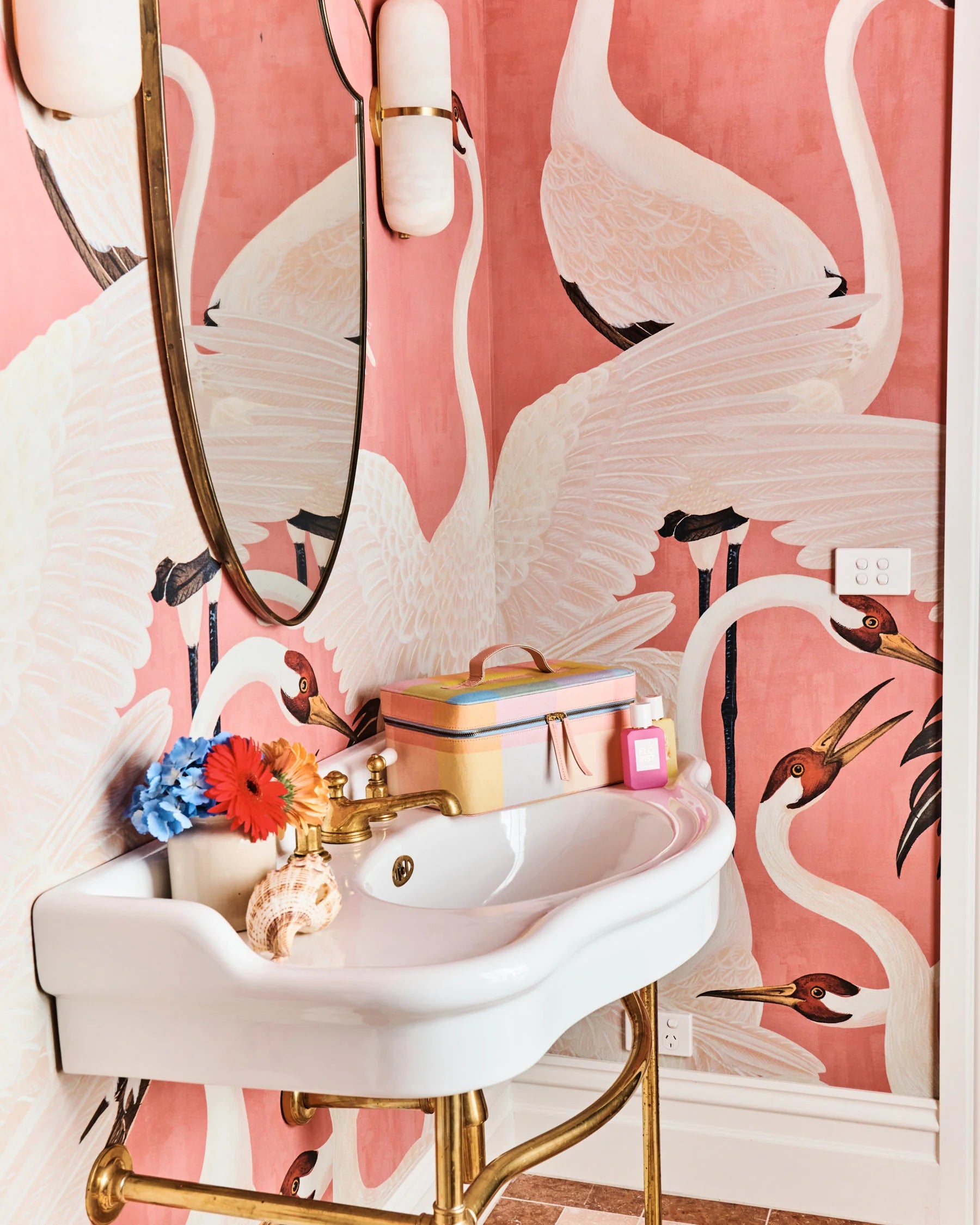 Corfu tartan pastel toiletry case sits on white vanity with gold legs. Skincare products and small vase with bright flowers sit on vanity. Gold mirror on pink bird wallpaper. 