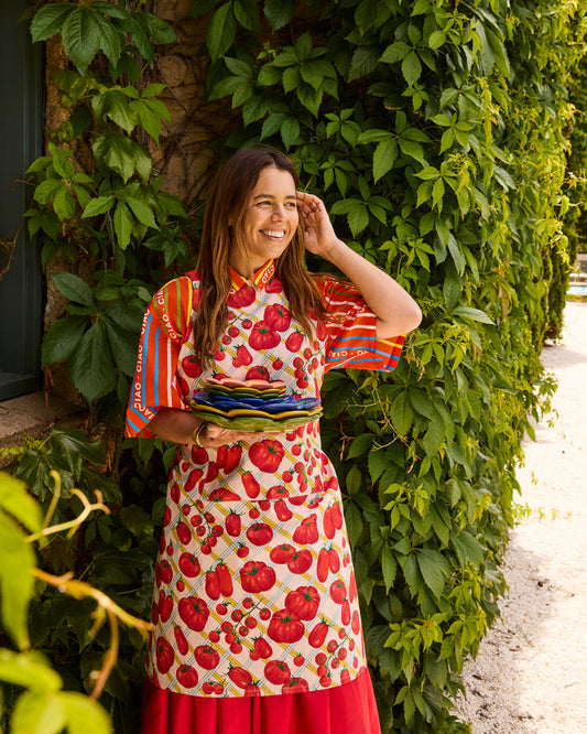 A woman stands outdoors against a wall covered in green ivy, wearing a colorful apron with a red tomato print over a striped shirt and red skirt, while holding a stack of plates.