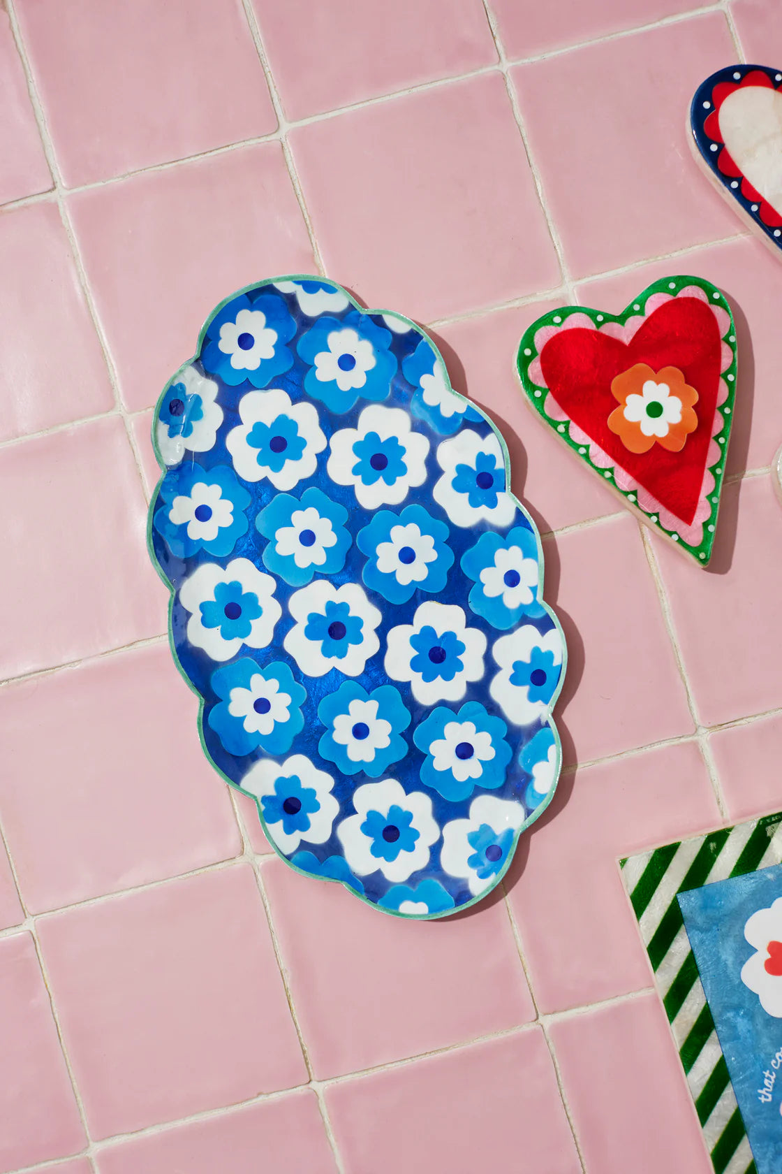 Floral-patterned blue sandal on a pink tiled floor with heart-shaped object and striped item in the background.