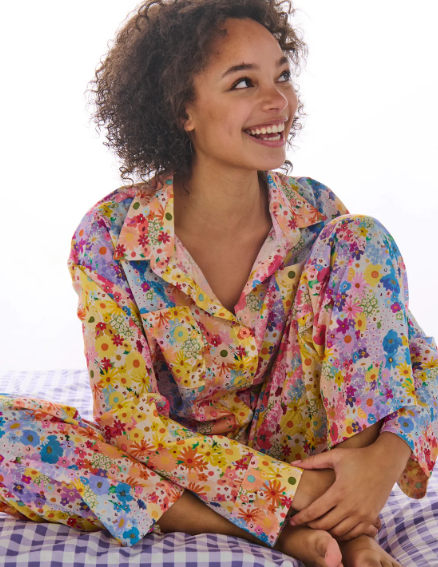 A smiling woman with curly hair sits cross-legged on a purple-and-white checkered bed, wearing a pastel floral pajama set with a multicolor print of flowers and butterflies.