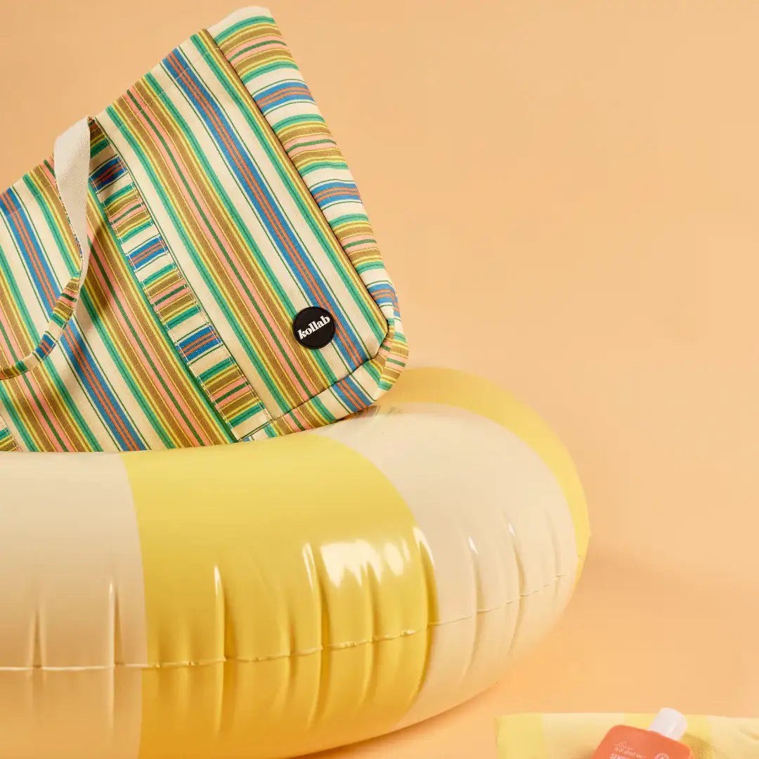 A striped tote bag with green, blue, yellow, and white vertical stripes, featuring a small black circular 'helio' logo, resting on a yellow and cream inflatable pool float against a solid peach background.