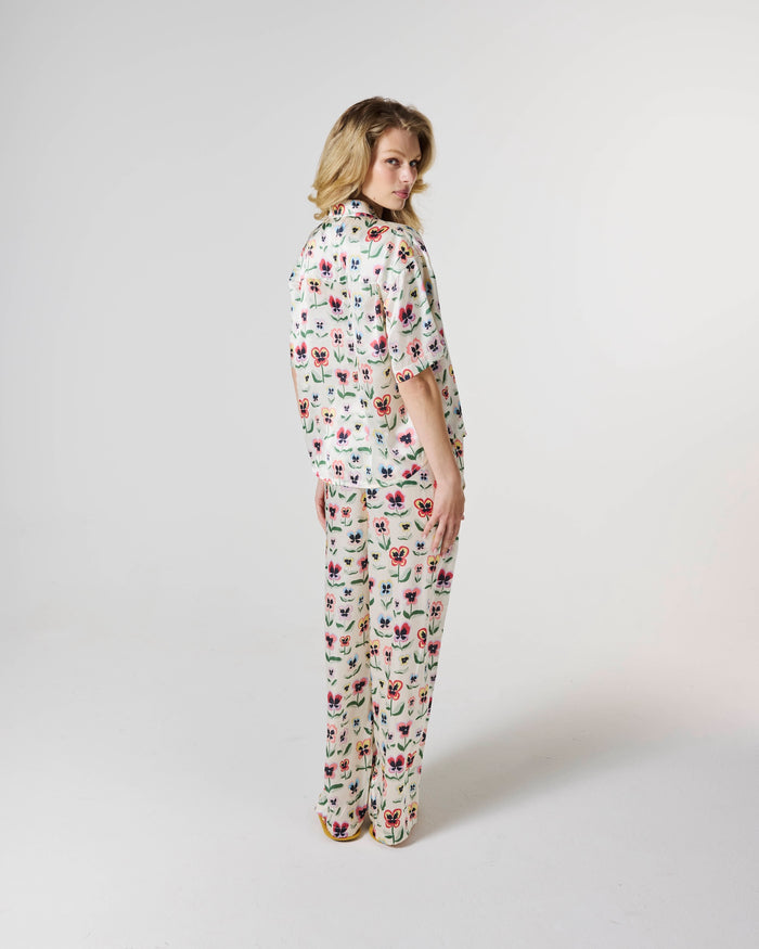 A woman modeling a matching two-piece pajama set with a short-sleeve button-up top and wide-leg pants, featuring an all-over floral print on a cream background. She is photographed from the back against a plain light gray studio backdrop.