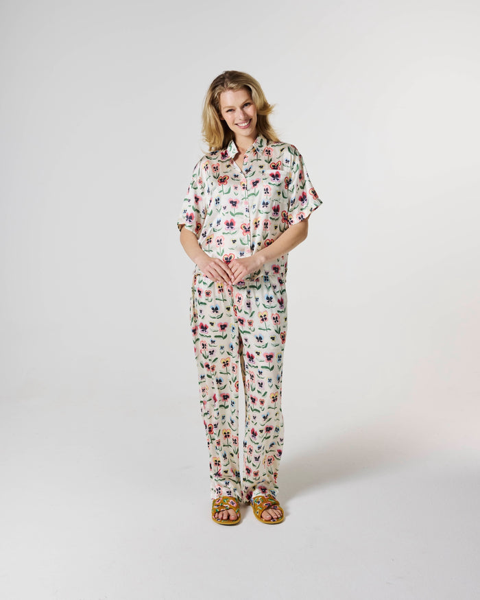 A woman modeling a matching short-sleeve button-up shirt and pajama pants set with a colorful floral print on a cream background, standing against a plain light gray studio background.