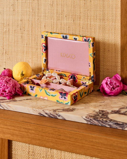 An open KIP&CO jewelry box with a yellow floral fabric exterior and a pink interior, displayed on a marble surface with flowers and a lemon against a burlap backdrop.