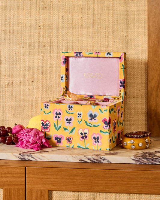 A yellow floral jewelry box with a pink interior lid, styled on a wooden table with decorative fruits and a small yellow container against a woven beige wall.