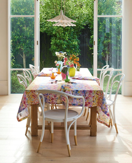 A colorful floral tablecloth with a matching napkin set on a wooden dining table, surrounded by white chairs in a bright room with large windows overlooking a green garden.