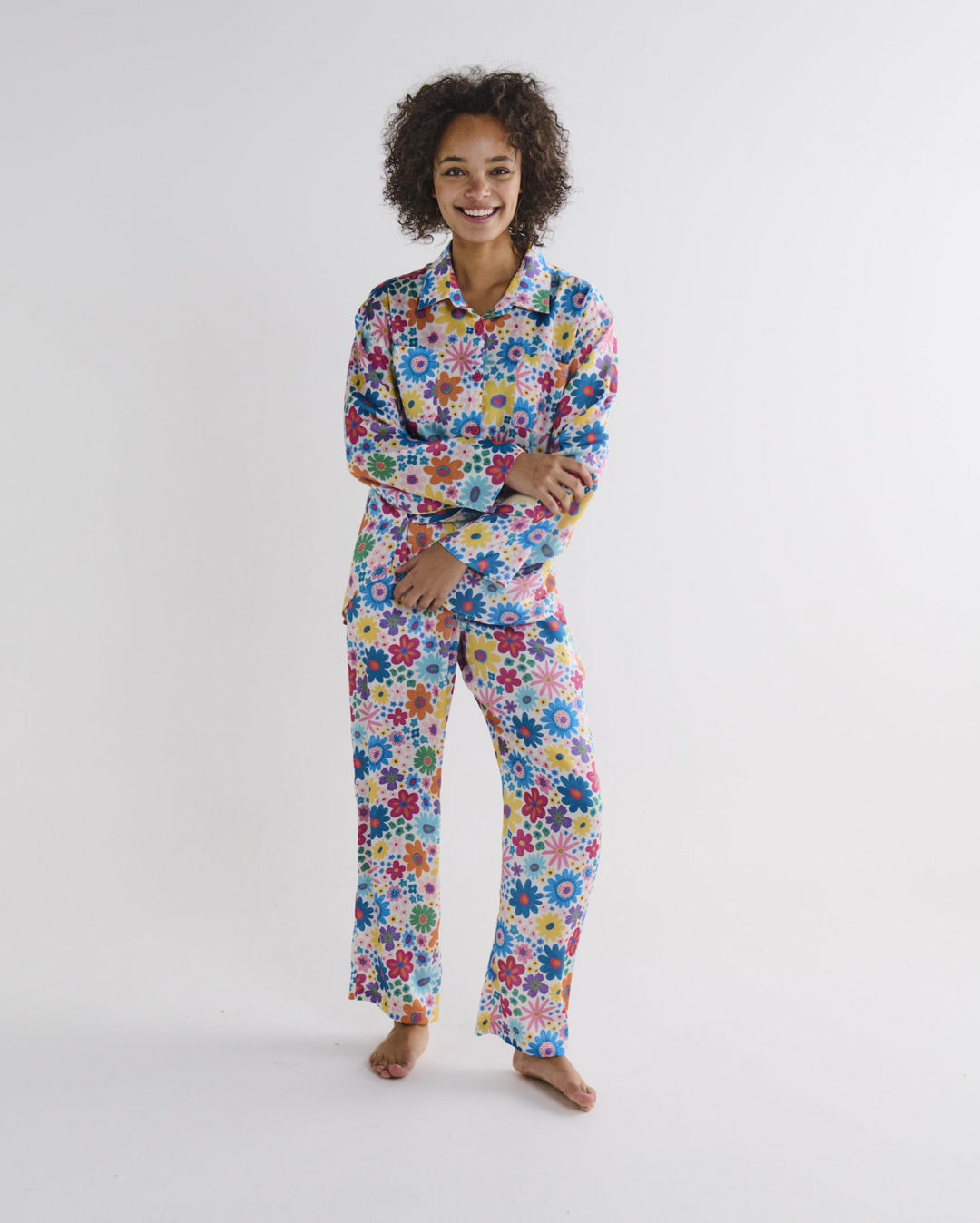 A woman modeling a two-piece pajama set with a colorful, dense floral pattern featuring blue, red, yellow, green, and pink flowers on a white background, photographed against a seamless light gray studio backdrop.