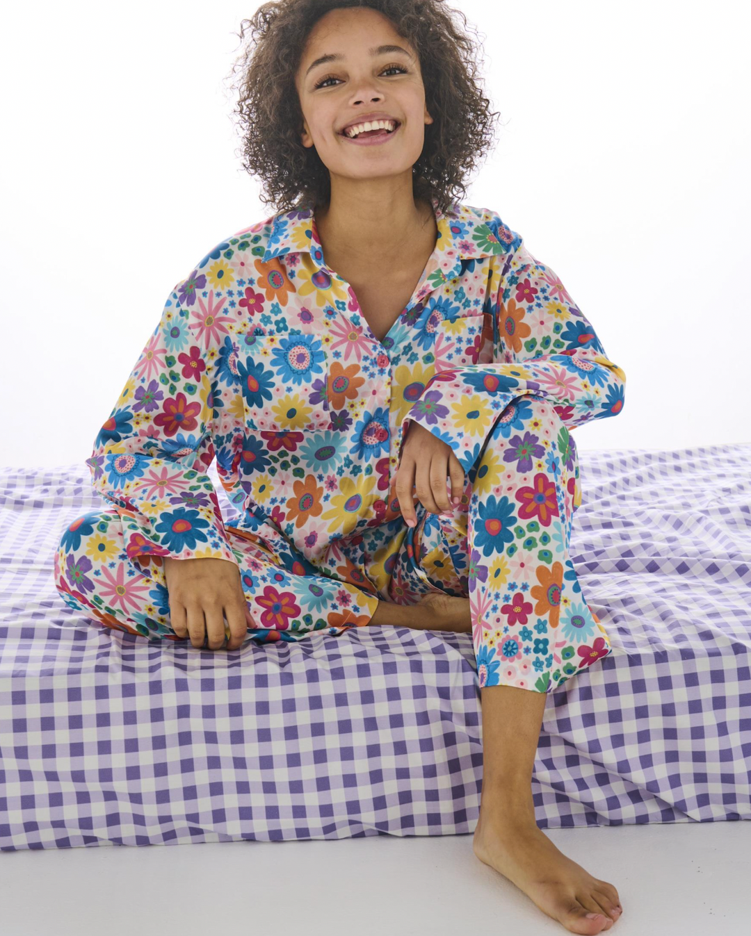 A smiling woman with curly hair sits cross-legged on a bed wearing a long-sleeve pajama set with a colorful floral print. The pajamas feature a mix of blue, red, yellow, green, and pink flowers on a light background.