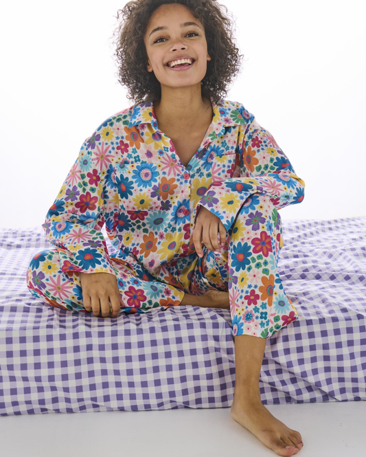 A smiling woman with curly hair sits cross-legged on a bed wearing a long-sleeve pajama set with a colorful floral print. The pajamas feature a mix of blue, red, yellow, green, and pink flowers on a light background.