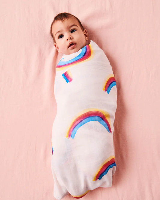 Baby wrapped in a white swaddle with rainbow patterns on a pink background