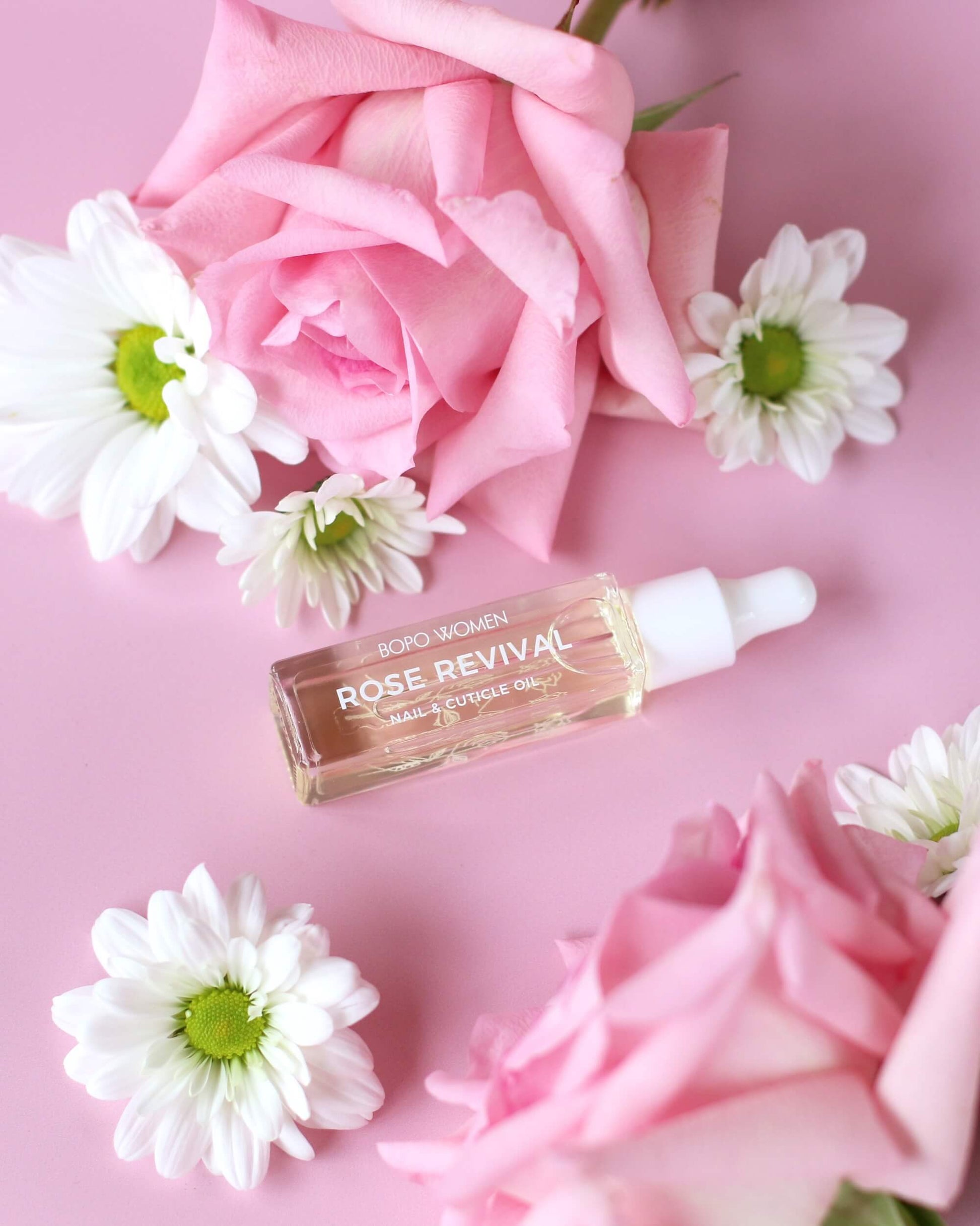Skincare nail and cuticle serum bottle labeled 'Rose Revival' surrounded by pink roses and white daisies on a pink background.