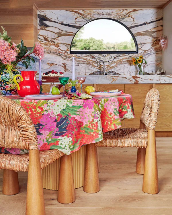 Bush blossom round tablecloth with chunky timber chairs sat around table, styled with red jug, colourful glassware, plates, cake stand and blossom flowers in vase. Marble counter top and wall in background with arch window. 